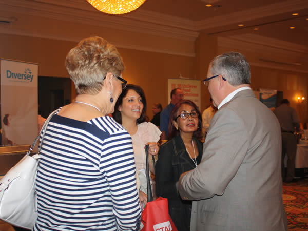 Four adults stand talking at an indoor event; one man in a suit faces three women, with event banners visible in the background.