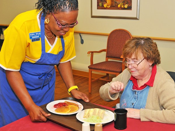 A caregiver in a yellow shirt serves a meal tray with pancakes and other food to an elderly woman seated at a table.