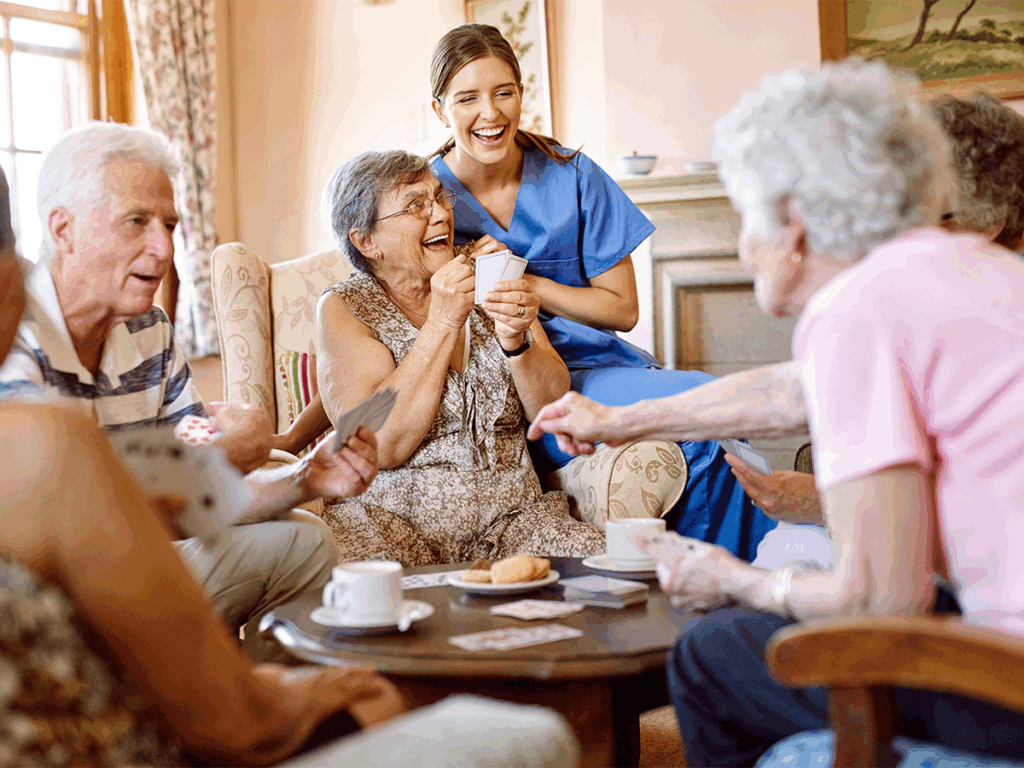 A group of seniors sit together playing cards while a caregiver in scrubs stands behind them, smiling, in a cozy living room setting.