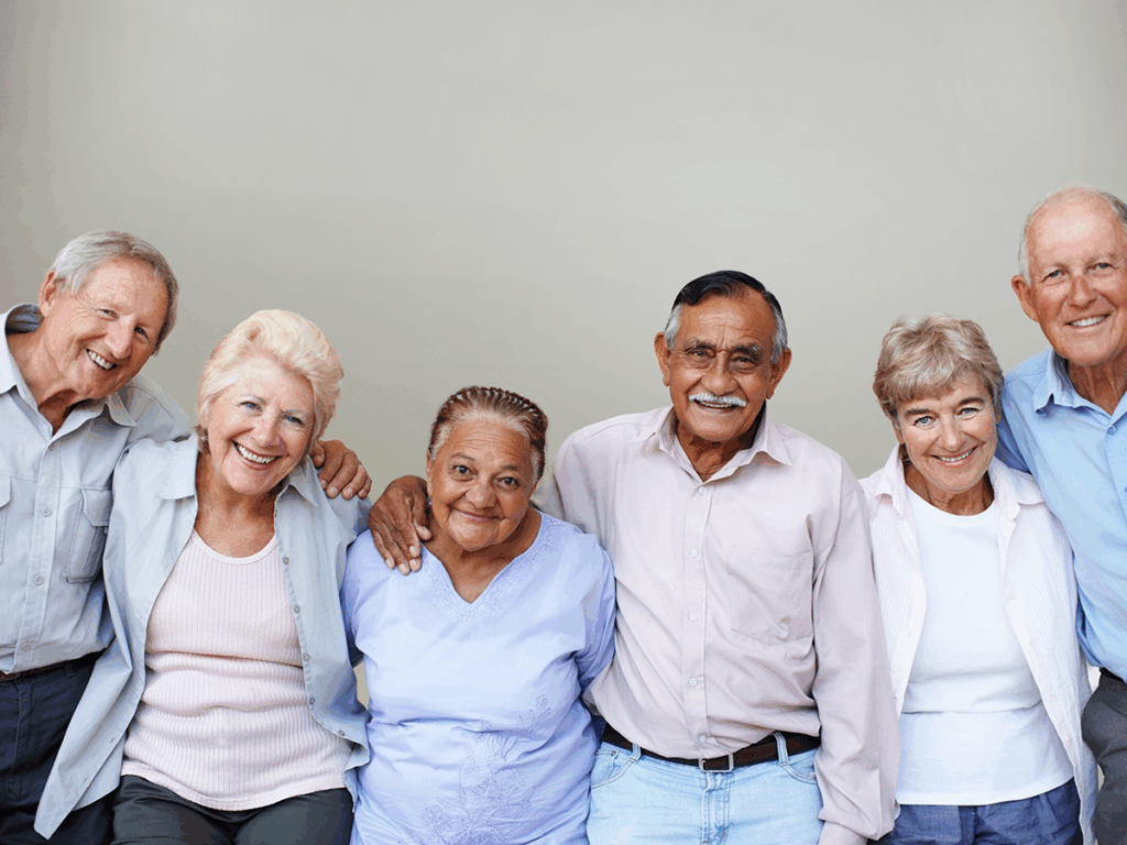 Six older adults stand closely together in a row, smiling at the camera against a plain light background.